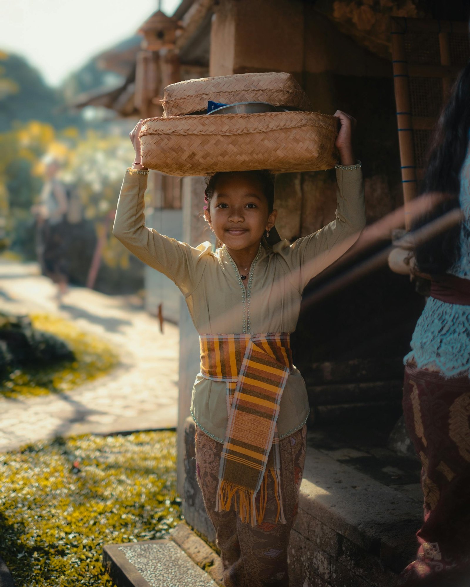 A young Balinese girl carries a traditional basket on her head in a sunny outdoor setting in Bali, Indonesia.