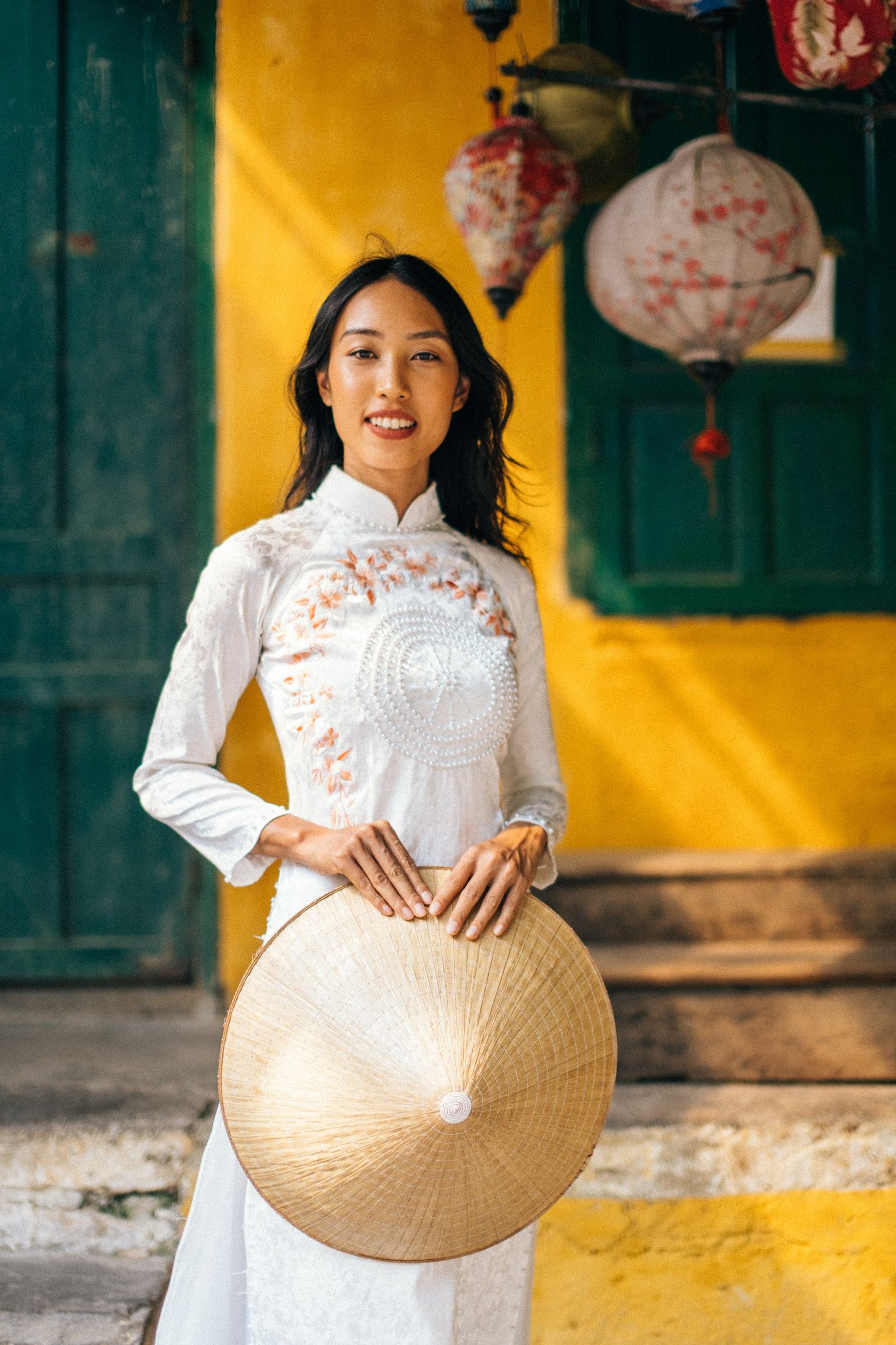 Charming woman in a white Ao Dai holding a conical hat, standing gracefully against a vibrant yellow wall.